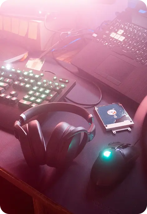 Modern tech workspace with laptop, mechanical keyboard, gaming mouse, over-ear headphones, and external hard drive on a desk with ambient RGB lighting.